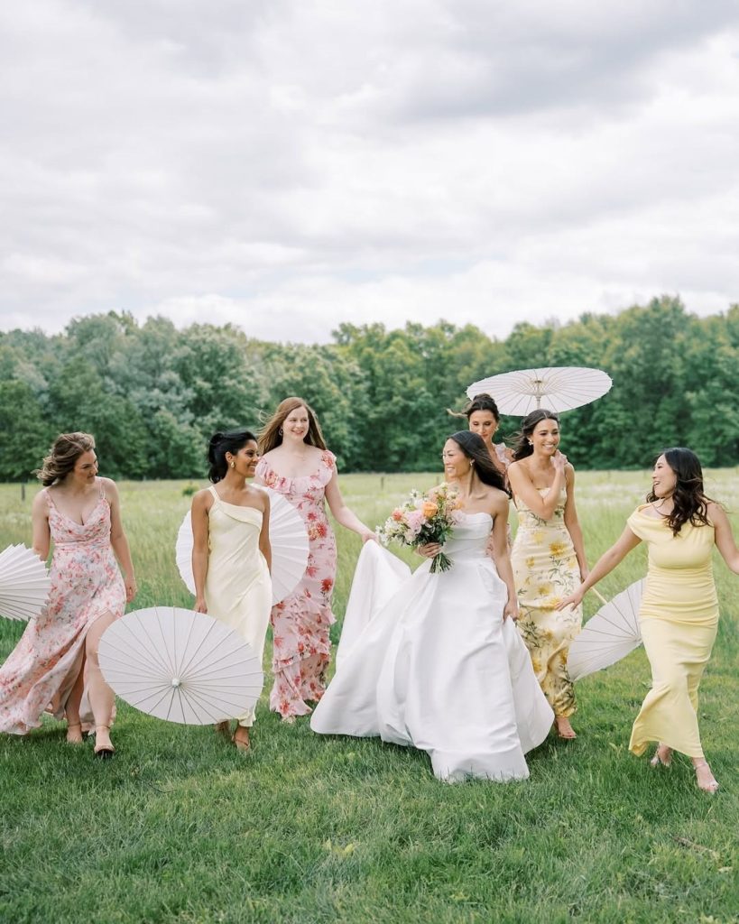 Bride holding a colorful bouquet walking through a grassy field with bridesmaids in pastel and floral dresses carrying white parasols.