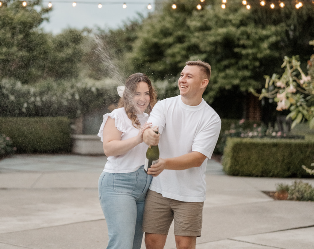 A smiling couple pops a champagne bottle during an outdoor engagement photo session, surrounded by lush greenery and glowing string lights.
