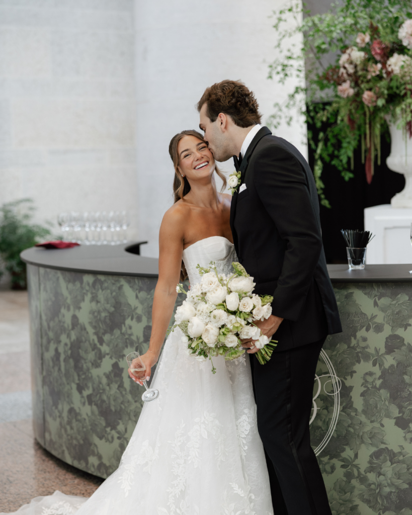 Groom Ben kisses bride Hannah on the cheek as she smiles and holds her white floral bouquet beside their custom monogrammed wedding bar.