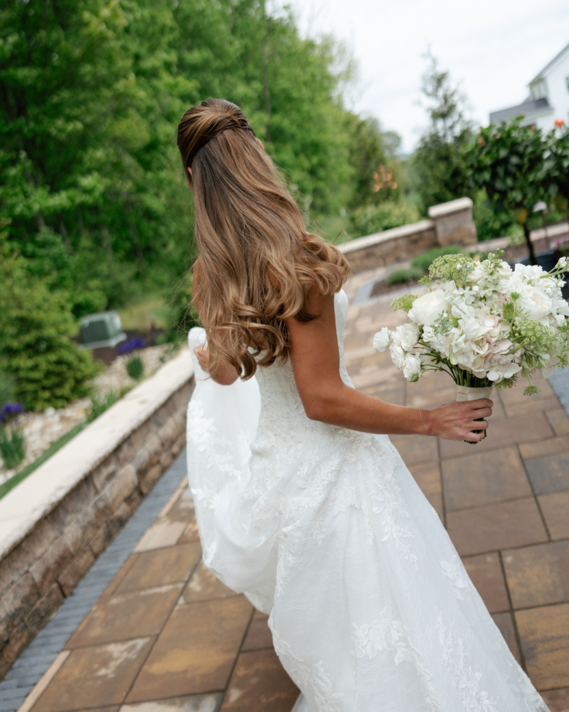Bride Hannah walks along an outdoor stone pathway carrying a white floral bouquet, showing the flowing lace detail of her wedding gown and curled half-up hairstyle.