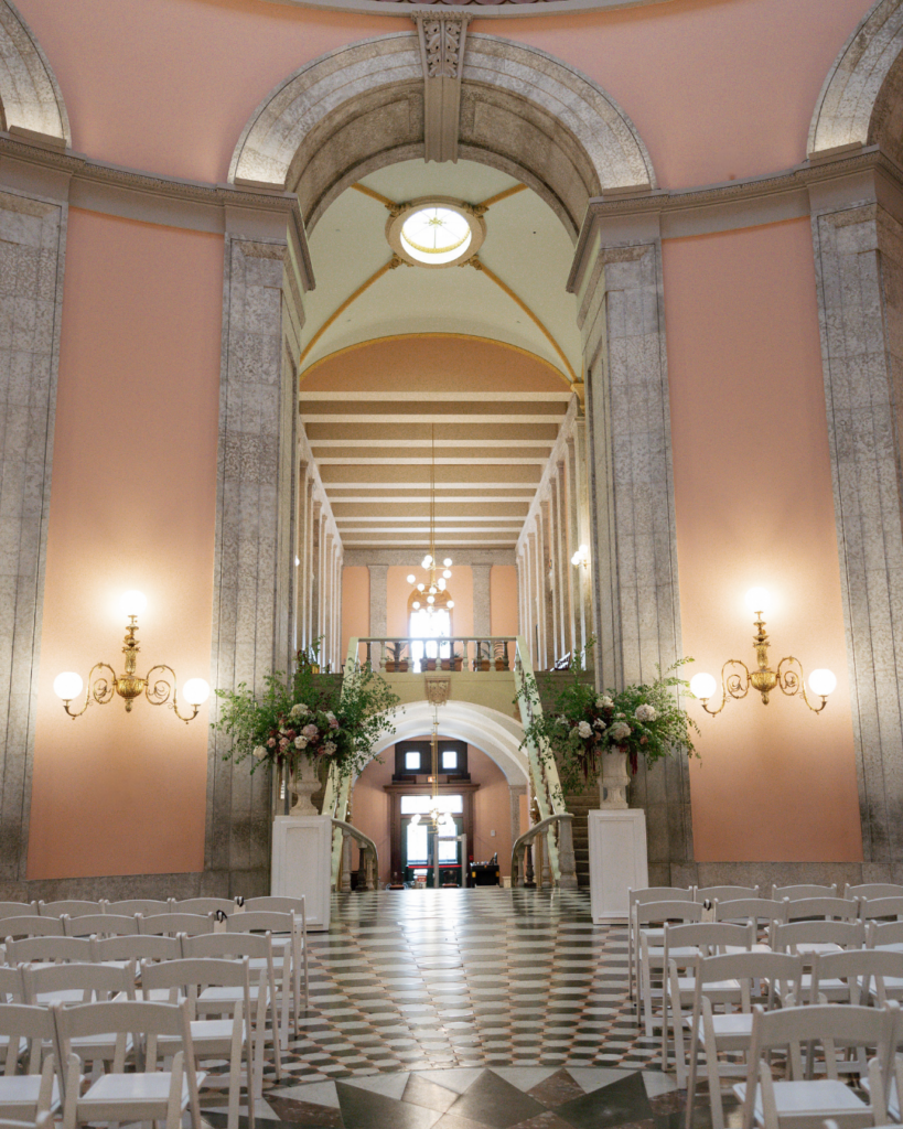 Wedding ceremony setup inside The Ohio Statehouse featuring white guest chairs, a black-and-white checkered aisle and large floral arrangements beneath grand arches.