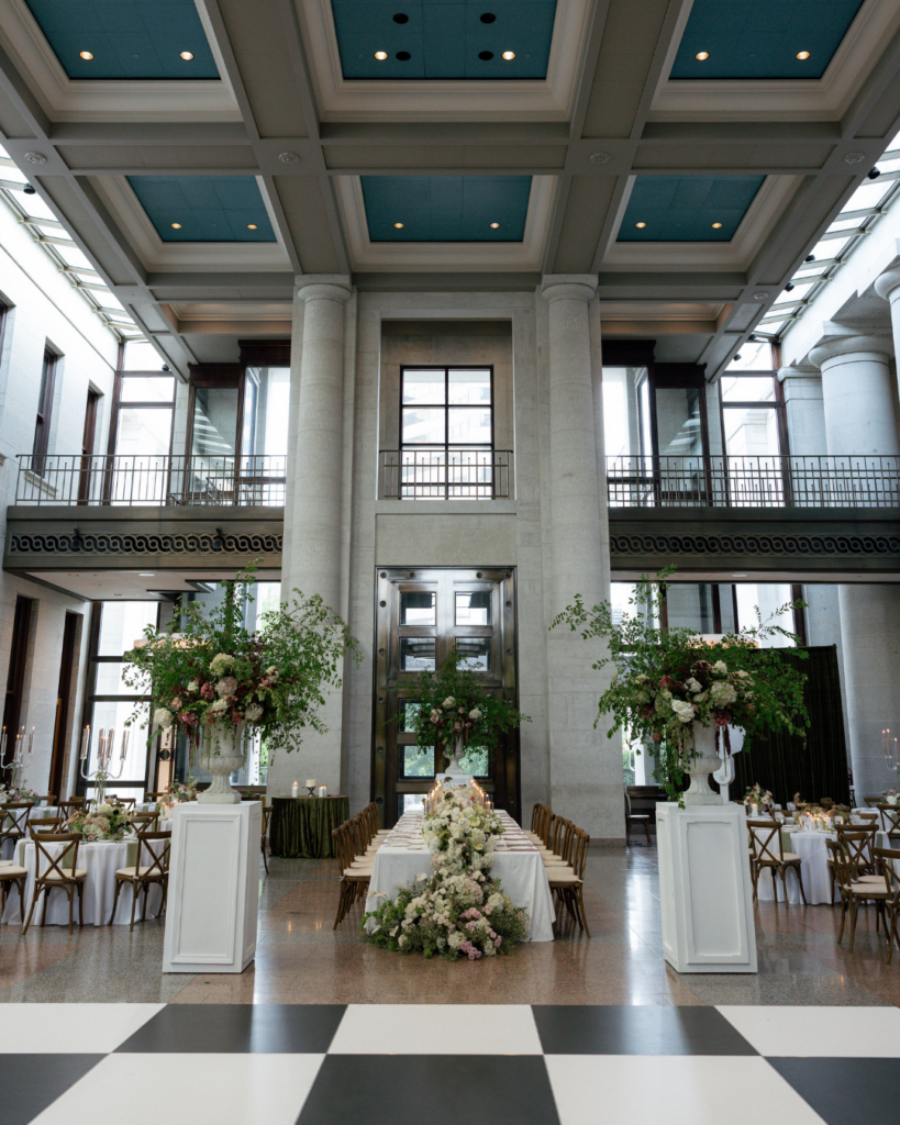 Grand Ohio Statehouse wedding reception with towering stone columns, lush floral urns, a long head table and a black-and-white checkered dance floor.