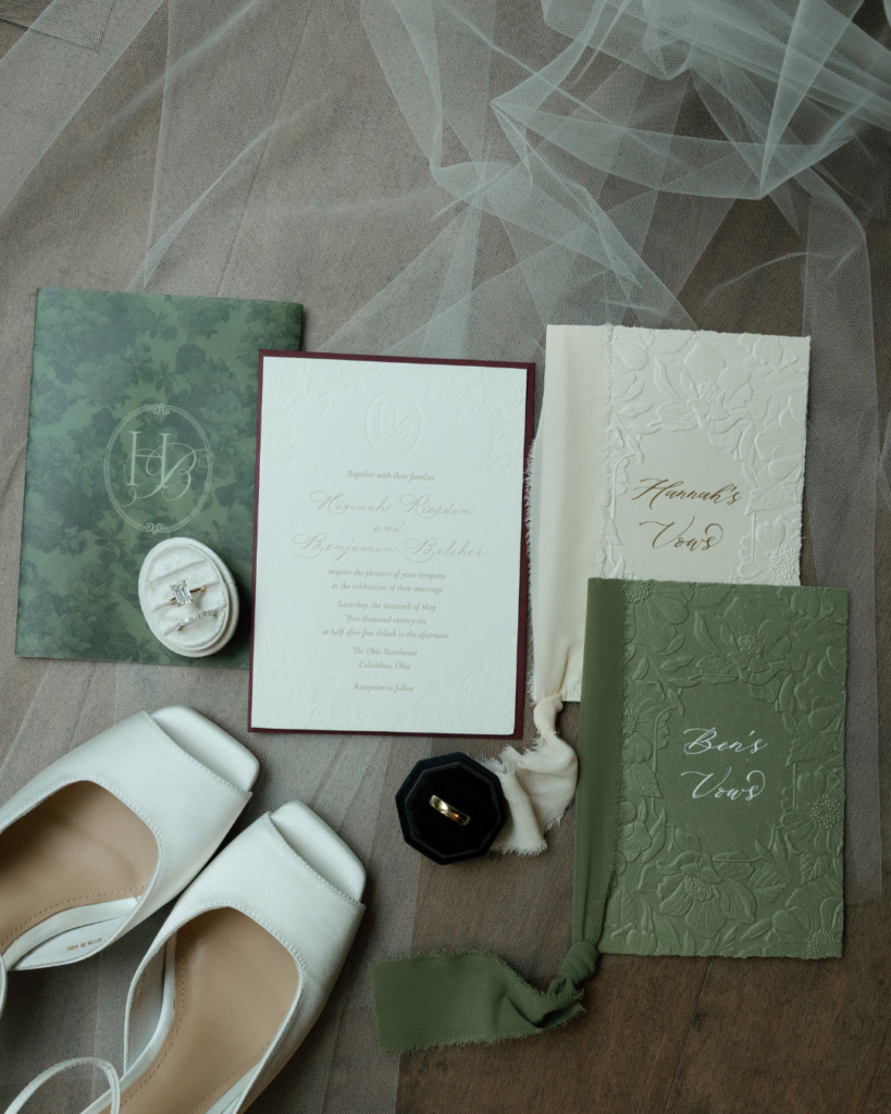 Custom wedding invitation suite by On Paper Press featuring Hannah and Ben's HB monogram, embossed stationery, sage green vow books, bridal shoes and wedding rings.