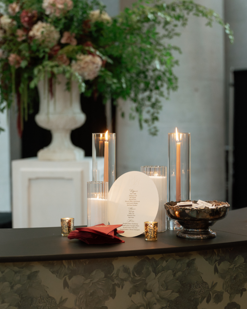 Romantic candlelit wedding bar details featuring glass hurricane candles, burgundy napkins, a custom oval drink menu and a floral-patterned bar front.