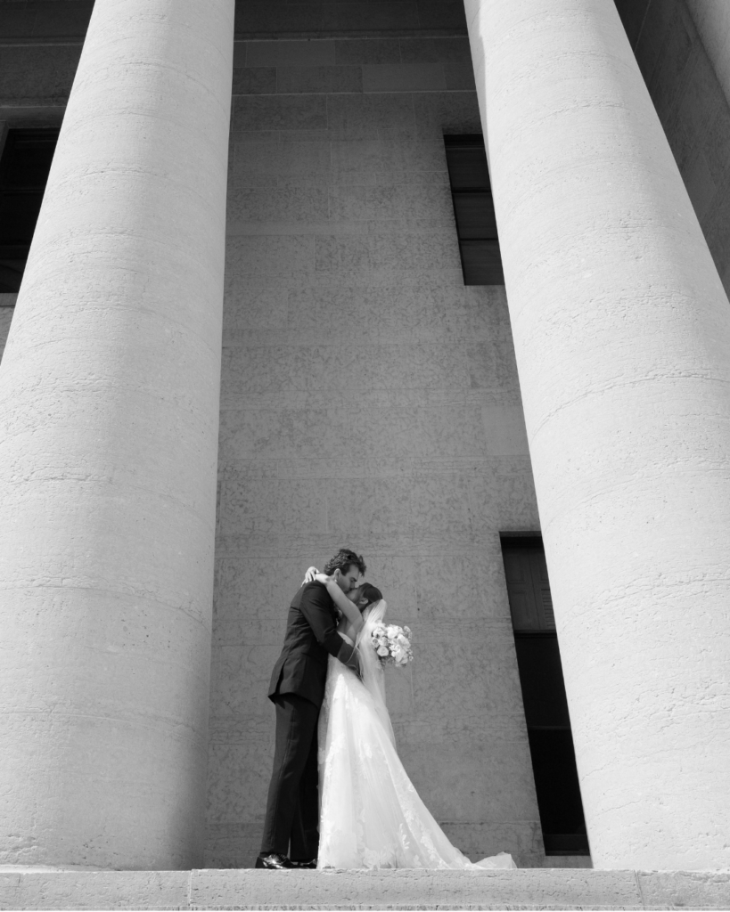 Black-and-white wedding portrait of Hannah and Ben kissing between the towering stone columns outside The Ohio Statehouse in Columbus, Ohio.