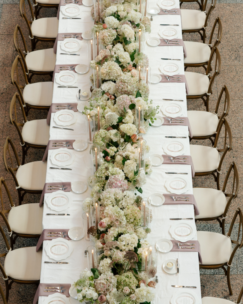 Overhead view of a romantic wedding head table with lush white and blush hydrangeas, roses, taper candles, dusty pink napkins and wooden cross-back chairs.