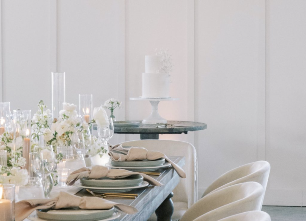 Two-tier white wedding cake with delicate floral detailing on a pedestal beside a candlelit reception table with white flowers and sage green plates.
