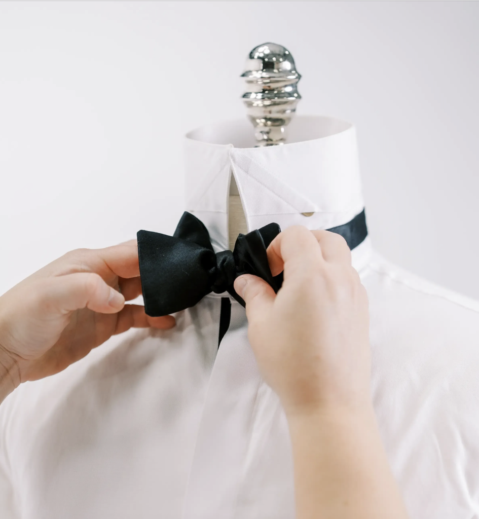 Hands adjusting a black bow tie on a white tuxedo shirt displayed on a mannequin, highlighting classic custom wedding suit styling in Columbus, Ohio.