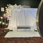 : Event Captura's elegant wedding photo booth backdrop featuring white draping, cascading blue and white florals, twinkling lights, glowing candles, and a gray velvet bench.