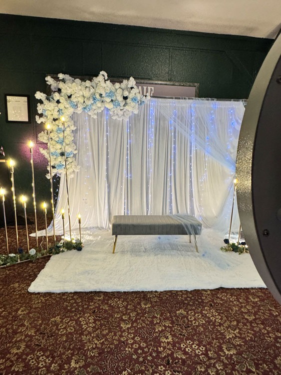 : Event Captura's elegant wedding photo booth backdrop featuring white draping, cascading blue and white florals, twinkling lights, glowing candles, and a gray velvet bench.