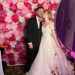 Bride kisses groom on the cheek while posing in front of a vibrant pink floral photo booth backdrop at a wedding celebration with Event Captura.