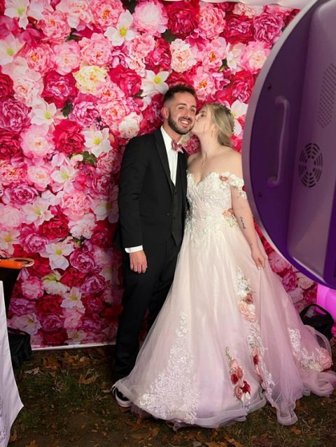Bride kisses groom on the cheek while posing in front of a vibrant pink floral photo booth backdrop at a wedding celebration with Event Captura.