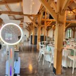 Event Captura's interactive wedding photo booth featuring a glowing circular LED light inside a rustic barn reception venue with wooden beams, white chairs, and floral centerpieces.