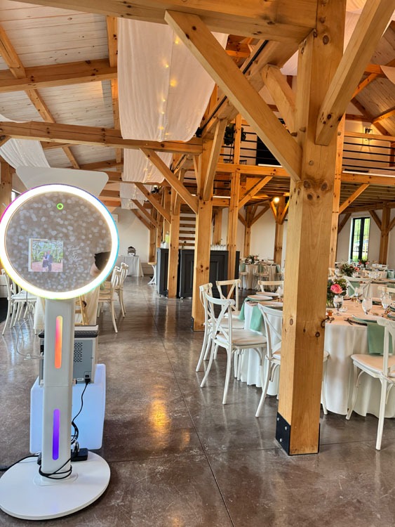 Event Captura's interactive wedding photo booth featuring a glowing circular LED light inside a rustic barn reception venue with wooden beams, white chairs, and floral centerpieces.