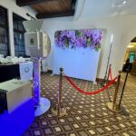 Event Captura's wedding photo booth setup featuring a purple floral backdrop, professional lighting, red velvet ropes, and a photo printer for guests to enjoy.