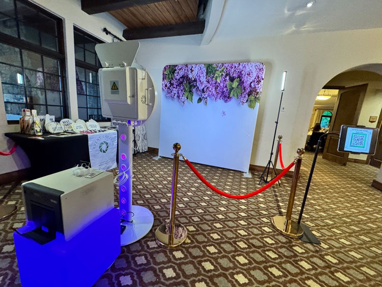 Event Captura's wedding photo booth setup featuring a purple floral backdrop, professional lighting, red velvet ropes, and a photo printer for guests to enjoy.