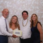 Newlyweds pose with two family members in front of Event Captura's soft white bokeh photo booth backdrop while holding a Happily Married sign.