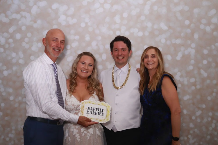 Newlyweds pose with two family members in front of Event Captura's soft white bokeh photo booth backdrop while holding a Happily Married sign.