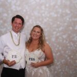 Bride and groom smile together at Event Captura's wedding photo booth, holding decorative Mr and Mrs signs in front of a sparkling white bokeh backdrop.