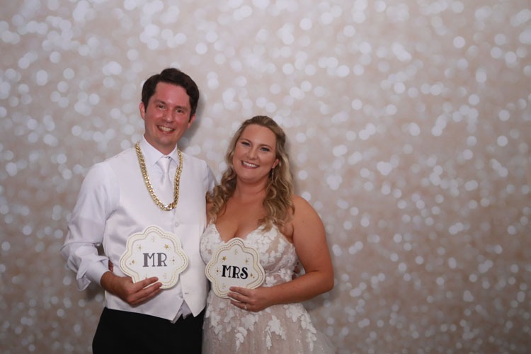 Bride and groom smile together at Event Captura's wedding photo booth, holding decorative Mr and Mrs signs in front of a sparkling white bokeh backdrop.