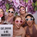 Four wedding guests pose playfully at Event Captura's photo booth with pink heart-shaped sunglasses, fun props, and an Eat, Drink & Be Married sign against a purple floral backdrop.