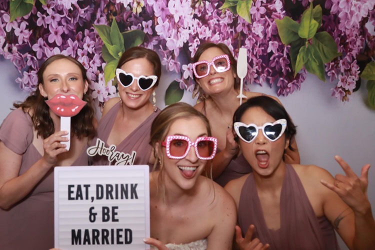 Four wedding guests pose playfully at Event Captura's photo booth with pink heart-shaped sunglasses, fun props, and an Eat, Drink & Be Married sign against a purple floral backdrop.
