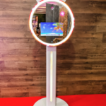 Event Captura's sleek white digital photo booth featuring a circular LED light, interactive touchscreen, and modern freestanding design against a wooden wall.