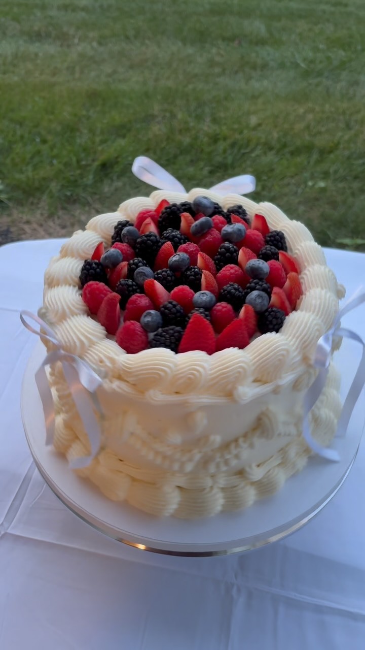 Helu Sweets Bakery custom vintage-style cake featuring intricate ivory buttercream piping, delicate white ribbon bows, and fresh strawberries, raspberries, blueberries, and blackberries.