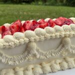 Helu Sweets Bakery rectangular vintage-inspired cake featuring elaborate ivory buttercream piping, decorative frosting swags, and fresh glazed strawberries displayed outdoors.