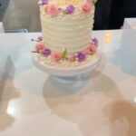 Helu Sweets Bakery custom white celebration cake featuring delicate textured buttercream frosting, pearl accents, and fresh pink roses and purple flowers arranged along the top and base.