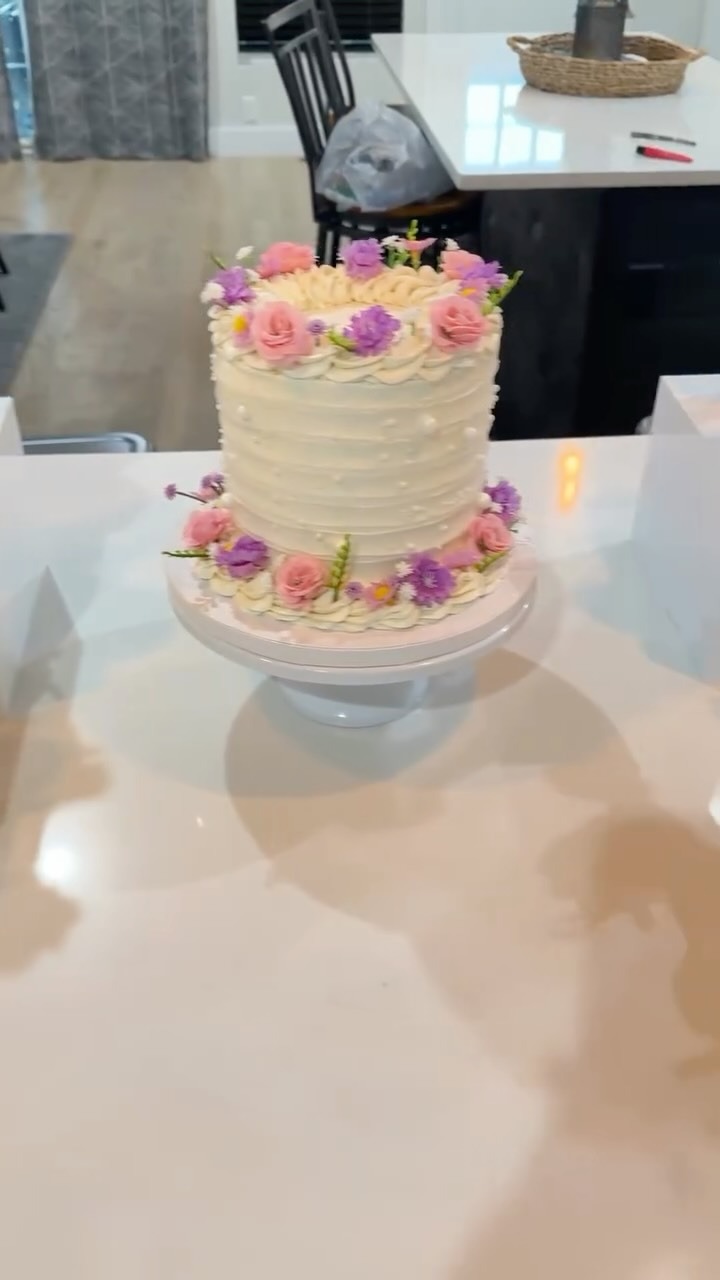 Helu Sweets Bakery custom white celebration cake featuring delicate textured buttercream frosting, pearl accents, and fresh pink roses and purple flowers arranged along the top and base.