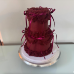 Helu Sweets Bakery two-tier burgundy cake featuring elaborate vintage-inspired buttercream piping, decorative frosting swags, and matching burgundy satin ribbon bows.