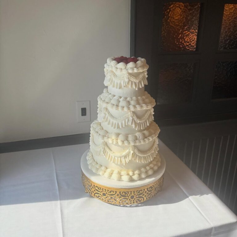 Helu Sweets Bakery elegant three-tier white wedding cake featuring intricate vintage Lambeth-style buttercream piping, cascading frosting swags, and decorative ruffles displayed on an ornate gold cake stand.