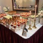 Helu Sweets Bakery wedding dessert table featuring strawberry cheesecake parfaits, layered chocolate desserts, decorated cupcakes, fresh floral arrangements, and gold candlesticks on a burgundy-draped table.