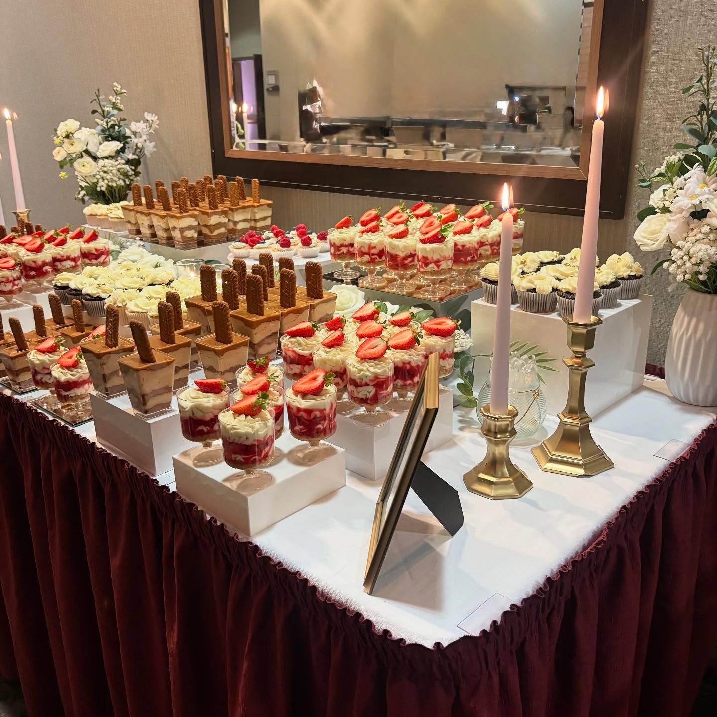 Helu Sweets Bakery wedding dessert table featuring strawberry cheesecake parfaits, layered chocolate desserts, decorated cupcakes, fresh floral arrangements, and gold candlesticks on a burgundy-draped table.