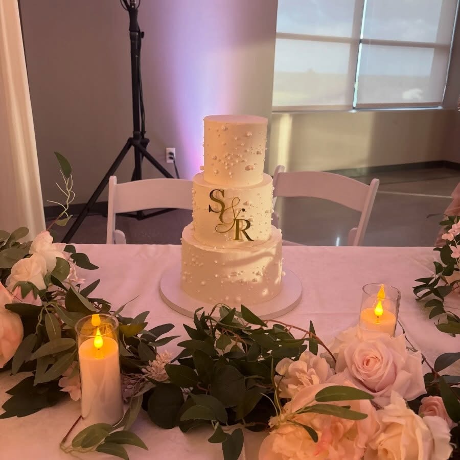 Elegant three-tier white wedding cake by Helu Sweets Bakery featuring delicate pearl accents, a gold monogram with the initials S and R, and a timeless minimalist design surrounded by romantic florals and candles.