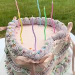 Helu Sweets Bakery pastel pink heart-shaped cake decorated with lavender and mint green buttercream piping, pink satin bows, colorful sprinkles, and whimsical multicolored birthday candles.