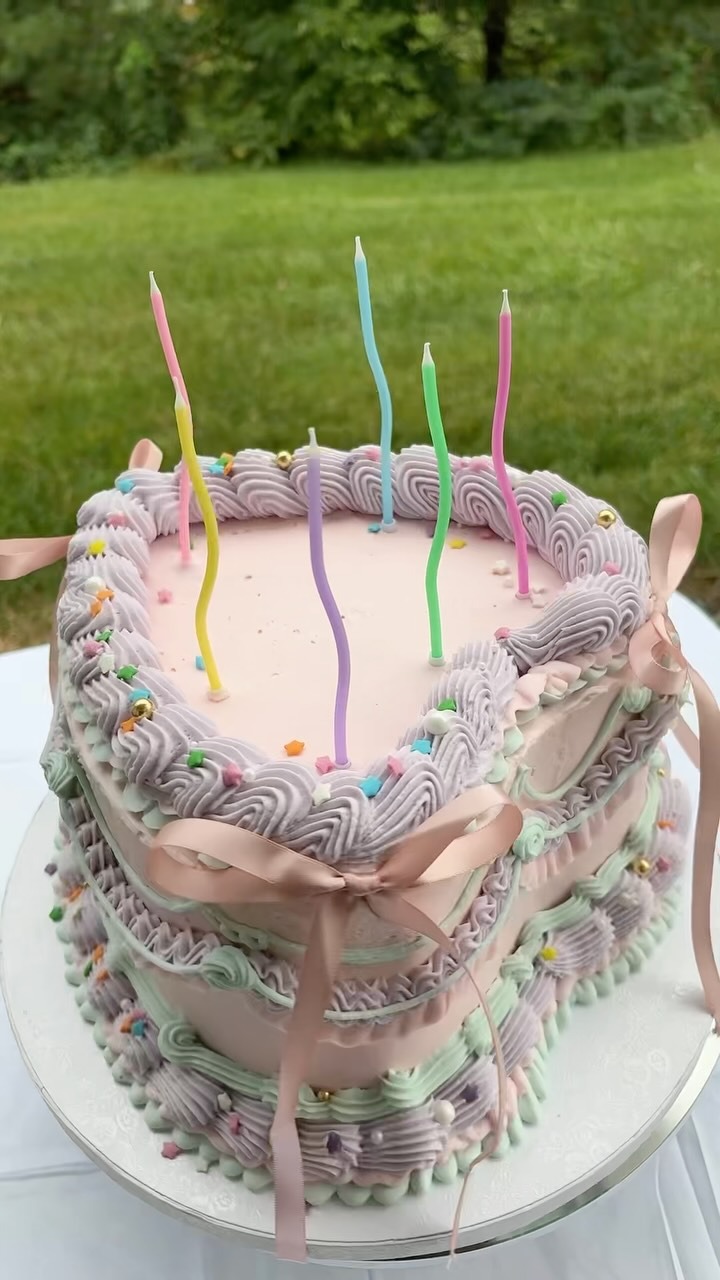 Helu Sweets Bakery pastel pink heart-shaped cake decorated with lavender and mint green buttercream piping, pink satin bows, colorful sprinkles, and whimsical multicolored birthday candles.