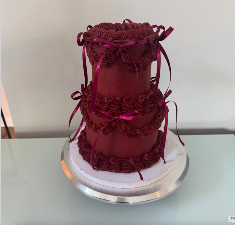 Helu Sweets Bakery two-tier burgundy cake featuring elaborate vintage-inspired buttercream piping, decorative frosting swags, and matching burgundy satin ribbon bows.
