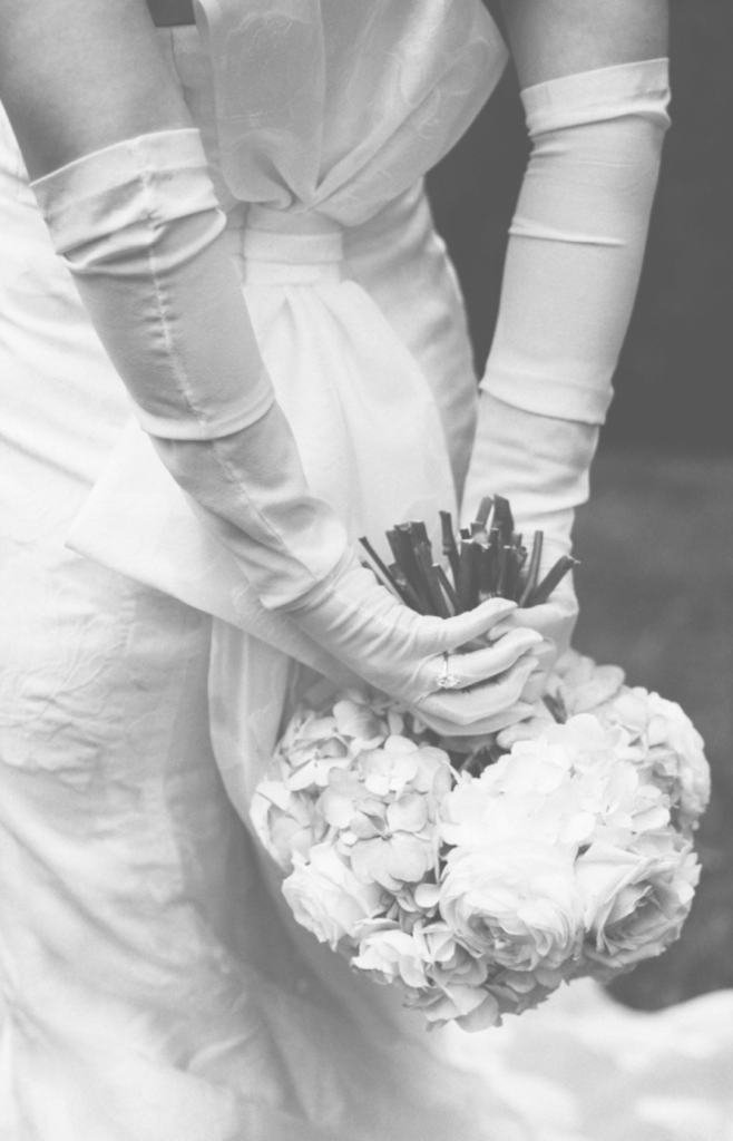 Bride in a tailored strapless gown with sheer white opera gloves, holding a large white wedding bouquet.