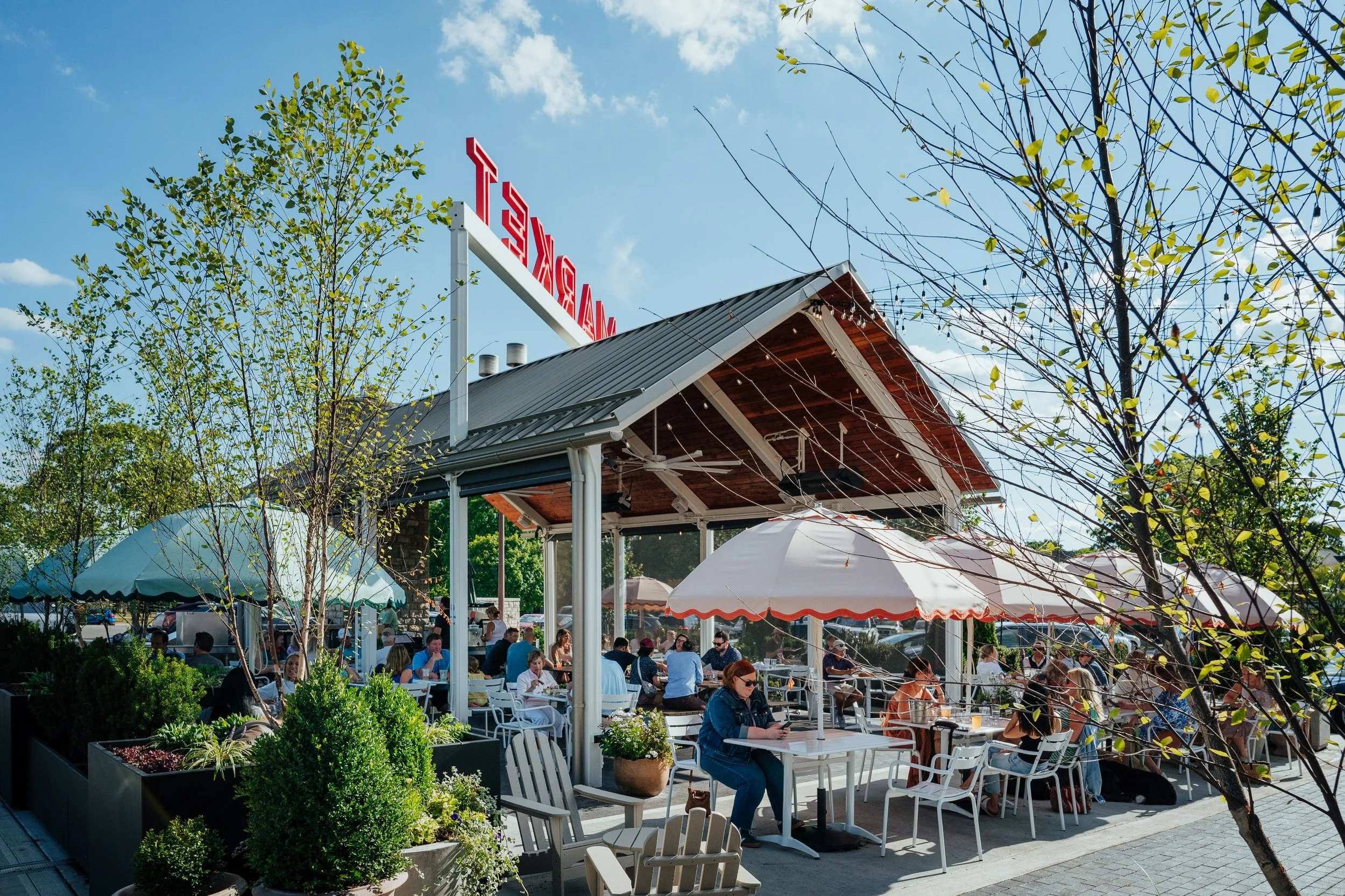 Outdoor patio dining area at Littleton’s Market in Upper Arlington, Ohio, featuring shaded tables, white umbrellas, greenery, and guests enjoying the sunny weather.