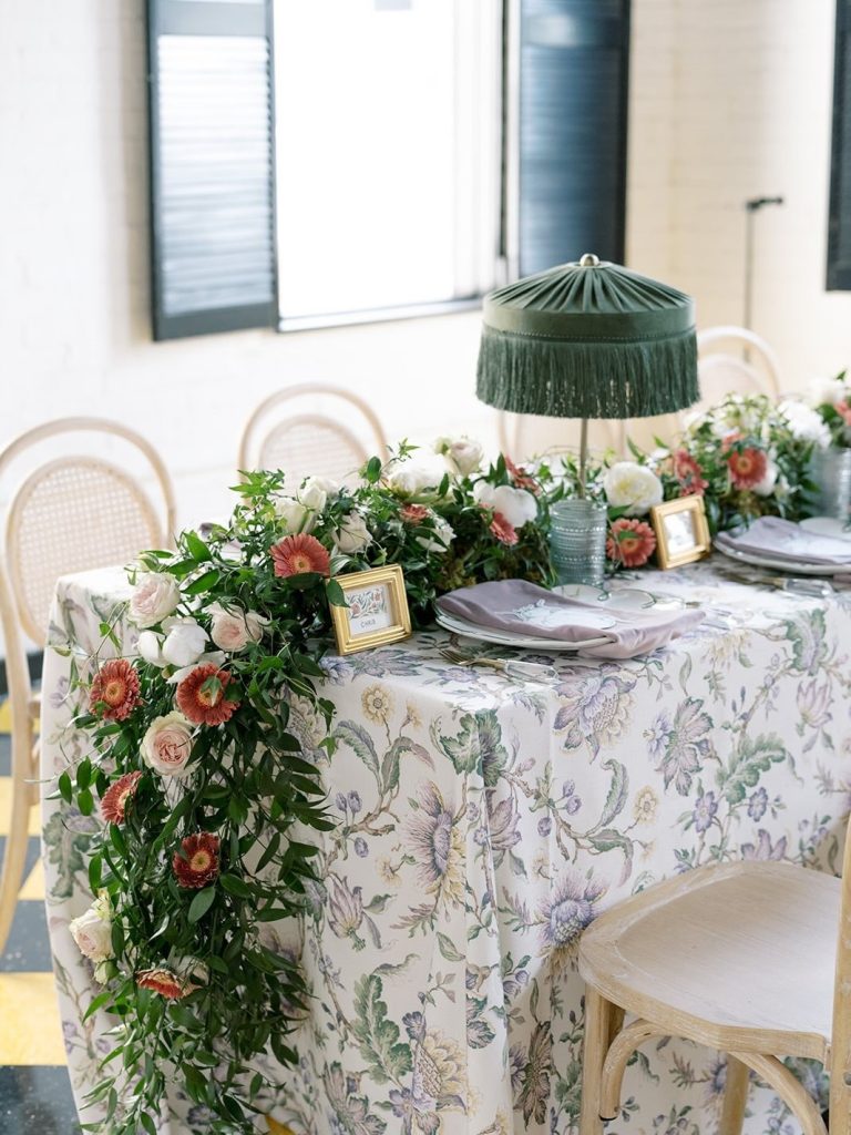 Romantic Columbus wedding reception tablescape featuring cascading pink and white florals by Market Blooms, patterned linens, green fringed lamps, and gold-framed place cards.