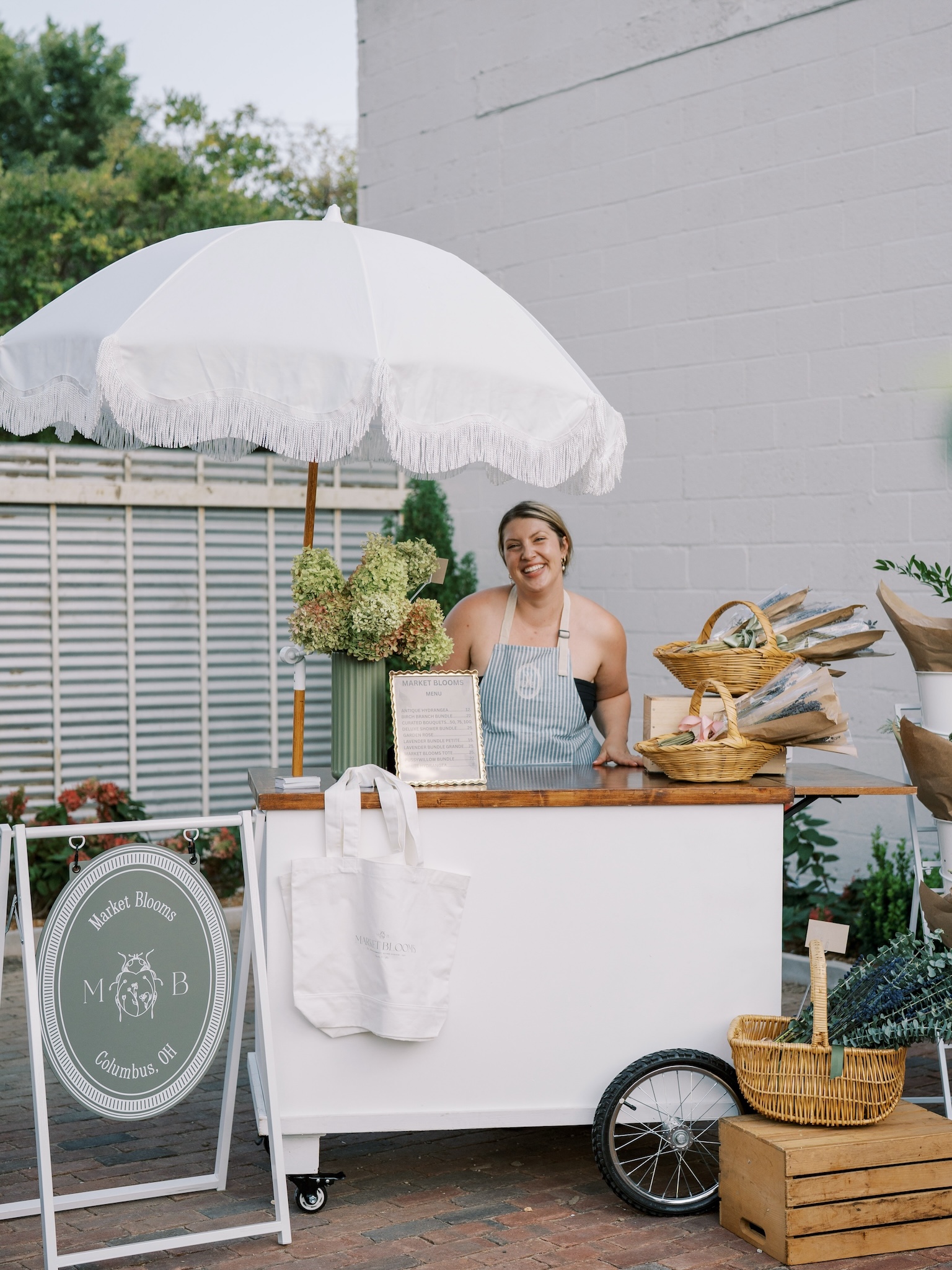 Market Blooms floral cart featuring fresh hydrangeas, wrapped bouquets, and a white fringed umbrella at an outdoor Columbus event.