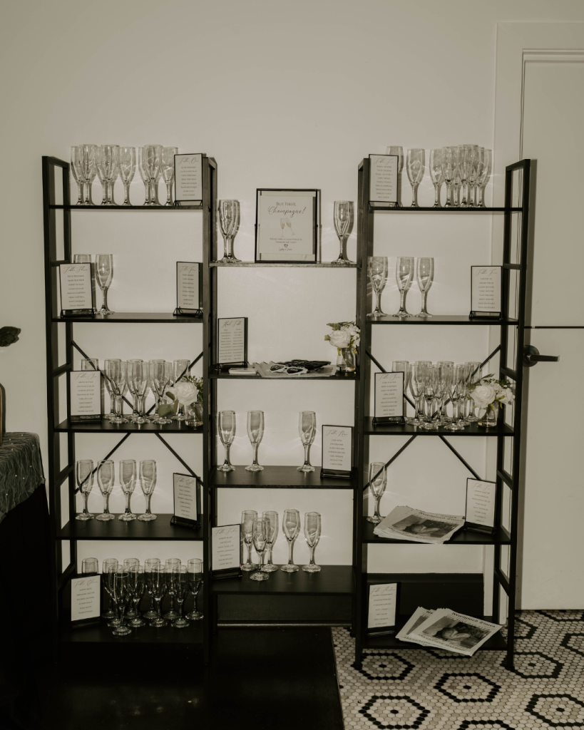Creative wedding champagne seating display featuring black shelving, champagne flutes, framed guest seating lists, and custom wedding newspapers at a Columbus reception.