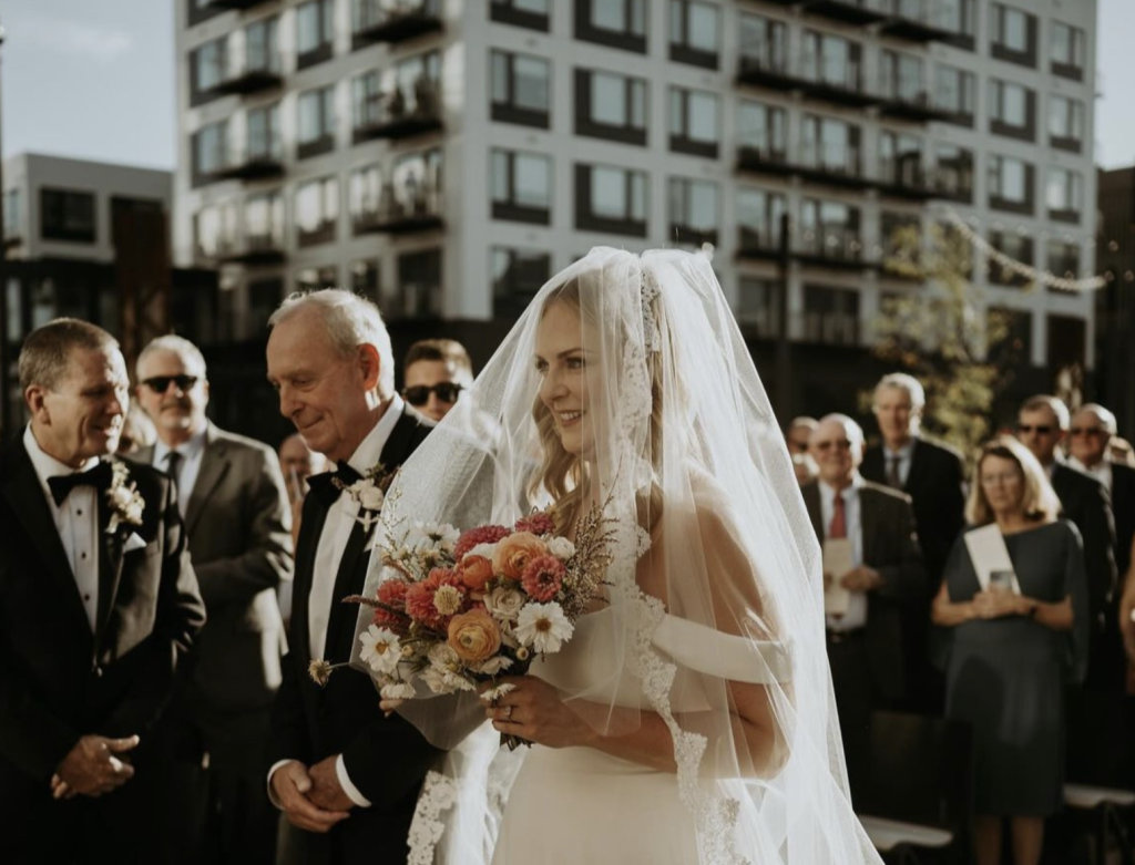 Bride holding a colorful orange, pink and white wedding bouquet while walking down the aisle with her father during an outdoor wedding ceremony in downtown Columbus, Ohio