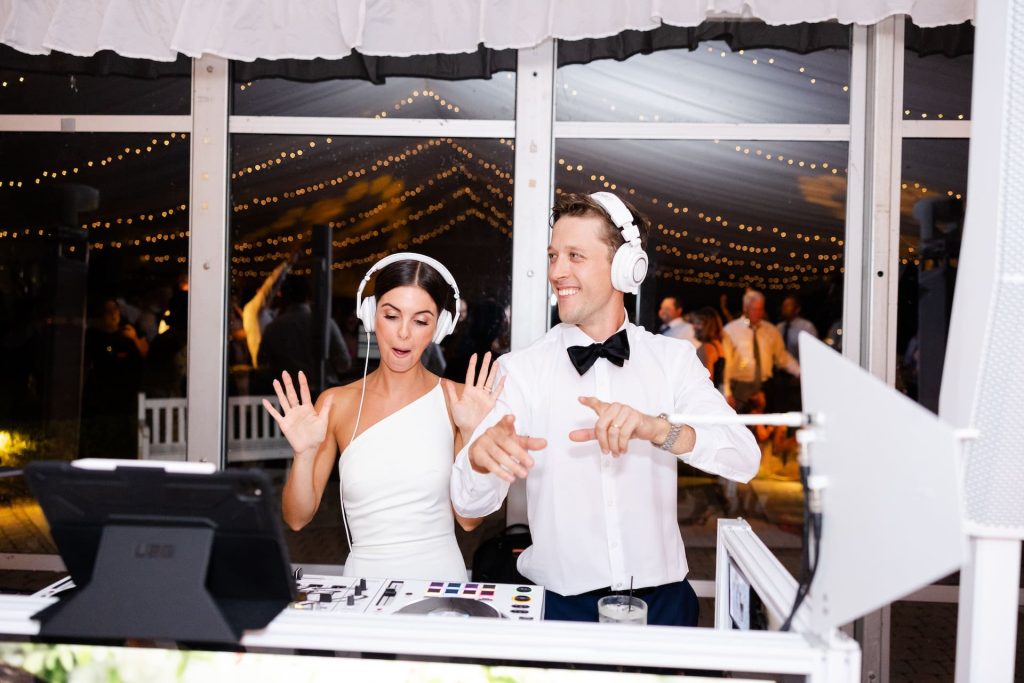 Columbus Ohio wedding DJ entertainment featuring a bride and groom wearing white headphones and mixing music at a DJ booth during their wedding reception, with glowing string lights and guests celebrating in the background.