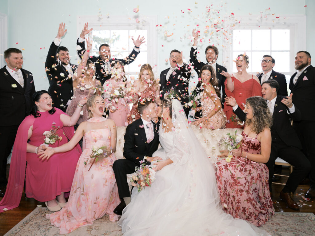 Bridgerton-inspired wedding party sitting indoors in front of large windows while rose petals are thrown in the air around them.