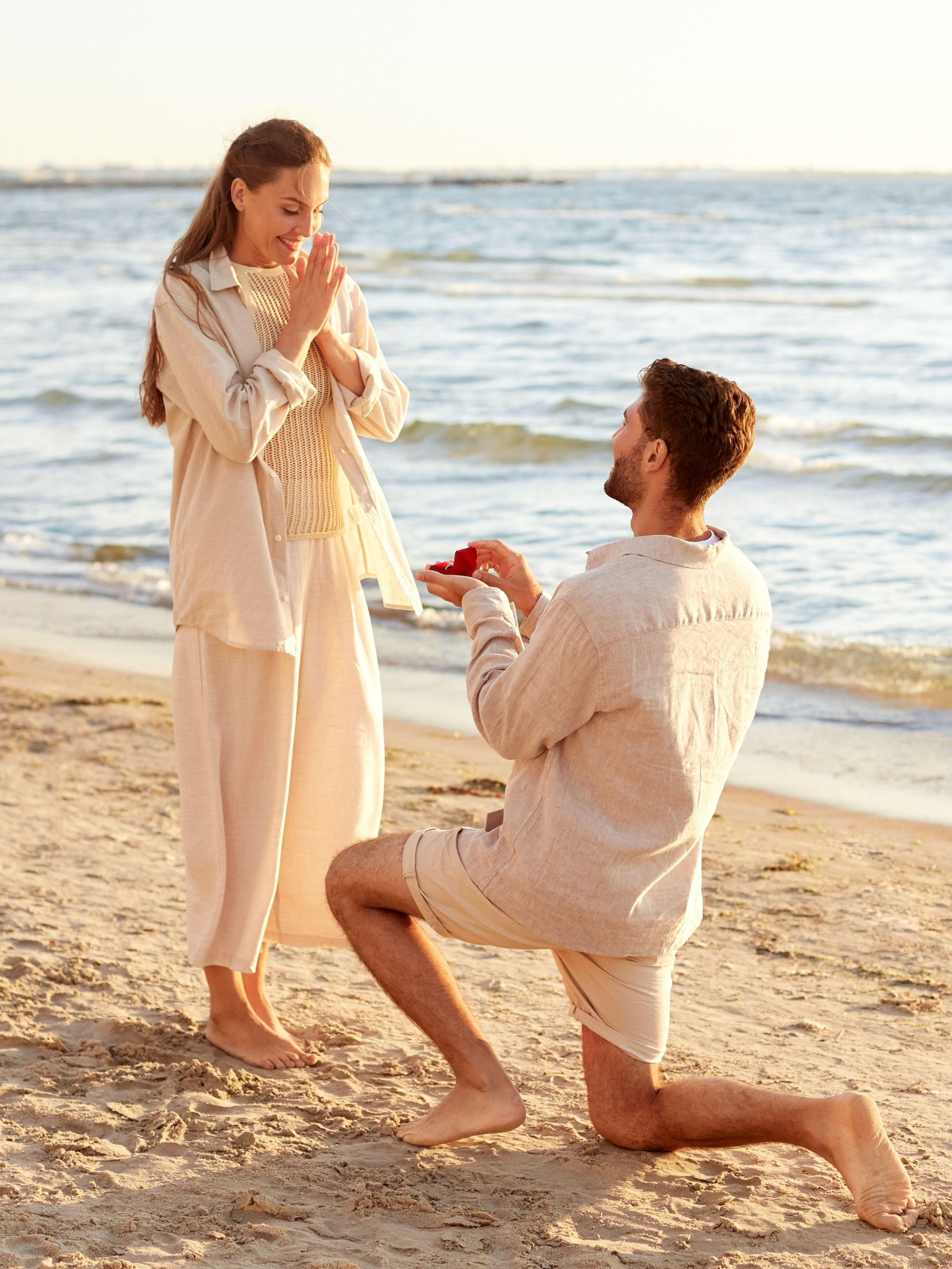 Man kneels on the beach holding a red ring box as he proposes to a smiling woman.