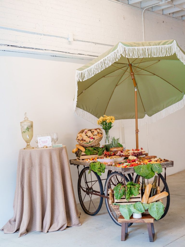 Wedding catering display featuring a rustic grazing cart with charcuterie, fresh fruit, bread, and appetizers beneath a green fringed umbrella, offering wedding catering inspiration in Columbus, Ohio.