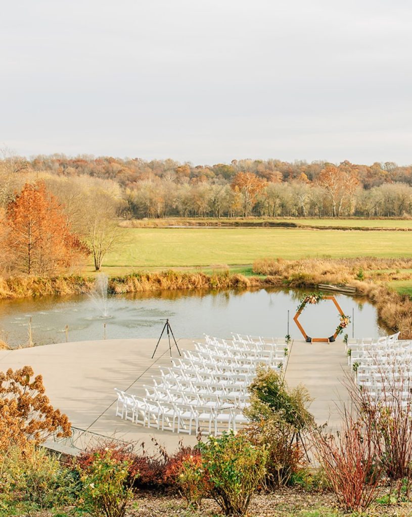 Outdoor wedding ceremony setup on a wooden deck overlooking a pond, featuring rows of white chairs, a geometric wooden arch, and colorful autumn foliage in the surrounding countryside.